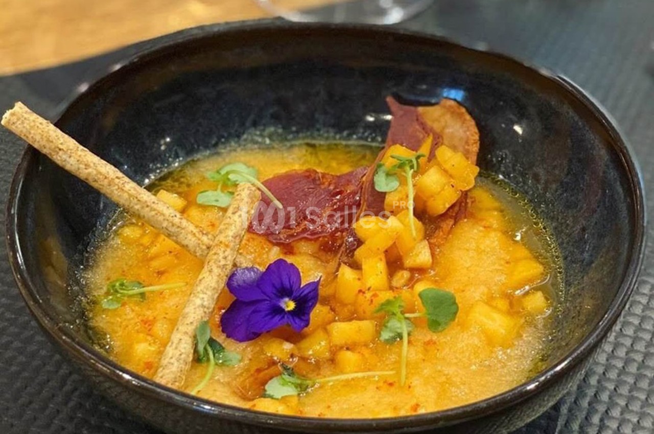 Soupe garnie de dés de légumes, chips de jambon, fleurs comestibles et gressins dans un bol noir.