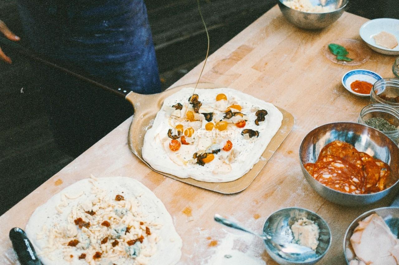 Préparation de pizzas maison sur une table en bois avec divers ingrédients comme tomates, fromage et charcuterie.