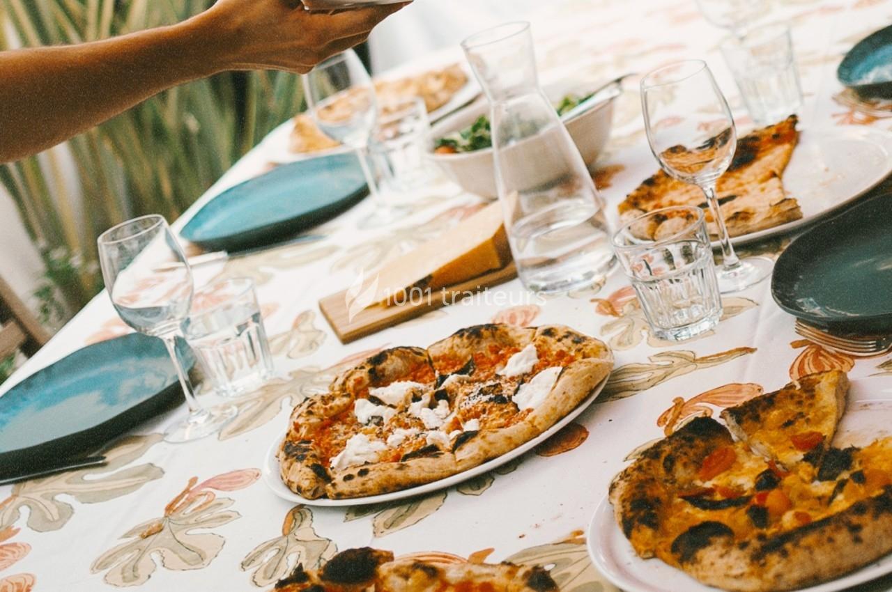 Table dressée avec des pizzas, assiettes vides, verres et carafe d'eau sur une nappe à motifs floraux.