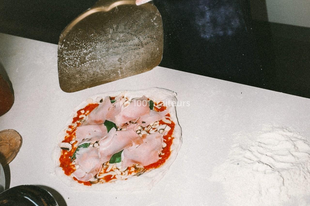 Pizza crue garnie de jambon et basilic sur un plan de travail, avec une pelle à pizza en arrière-plan.