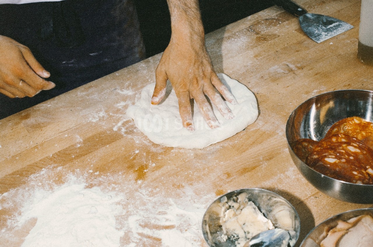 Une main aplatie une pâte à pizza sur une table en bois, entourée d'ingrédients et de farine.