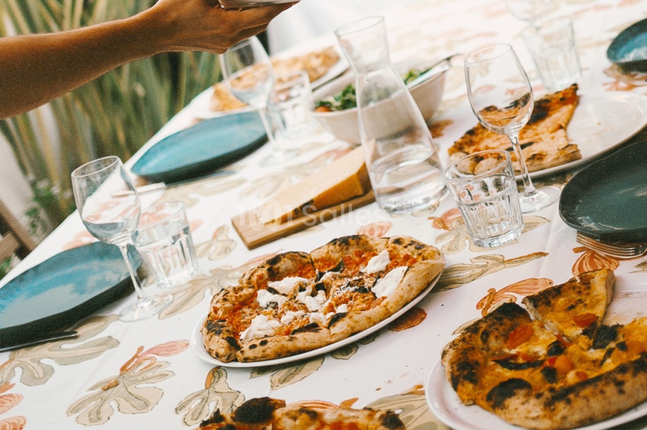 Table dressée avec des pizzas, assiettes vides, verres et carafe d'eau sur une nappe à motifs floraux.