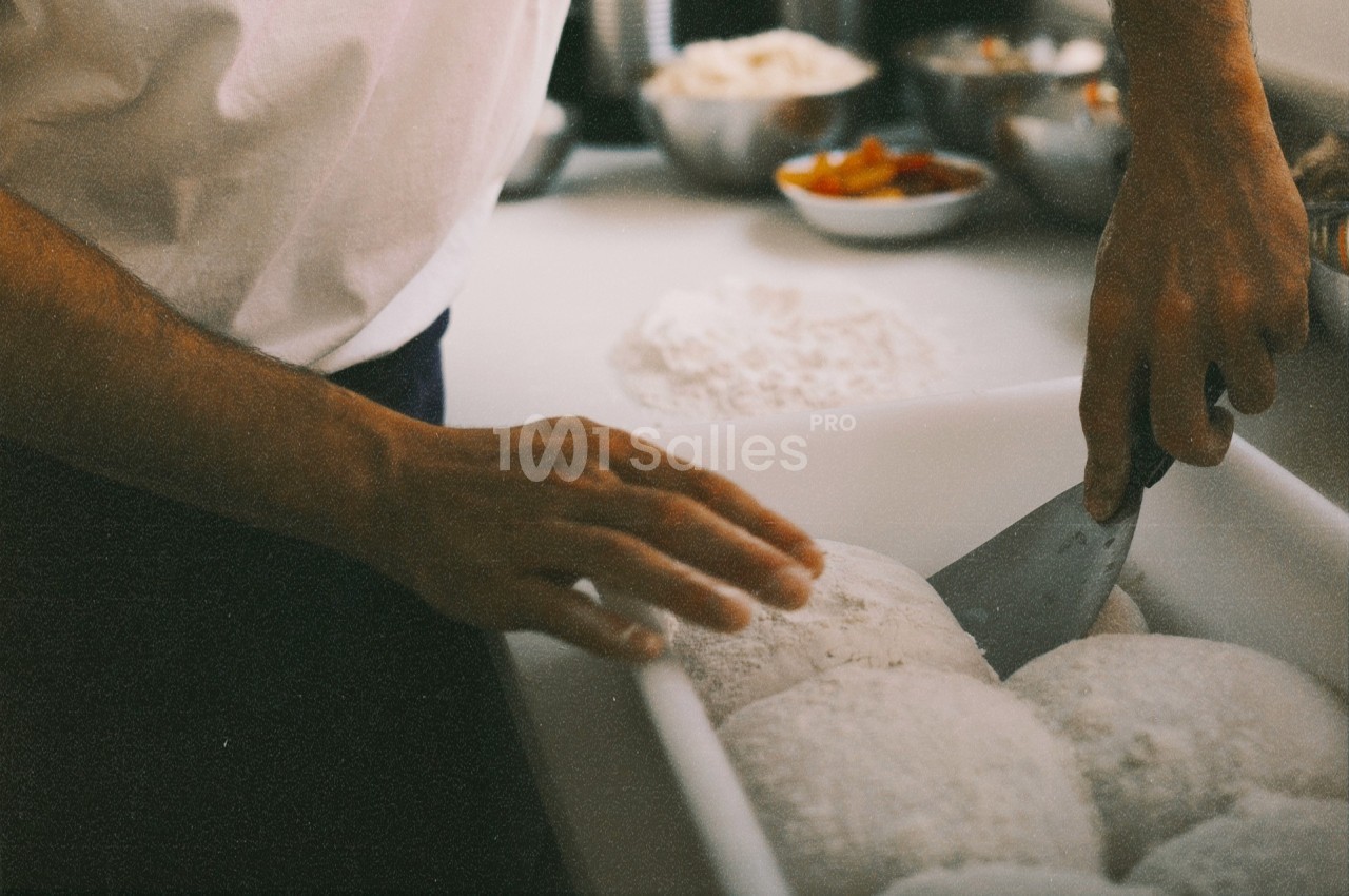 Un cuisinier façonne des pâtons de pâte sur un plan de travail avec un coupe-pâte, entouré d'ingrédients.