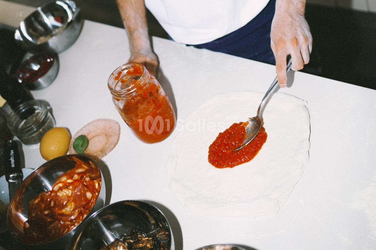 Une personne étale de la sauce tomate sur une pâte à pizza, entourée d'ingrédients dans des bols sur un plan de travail.