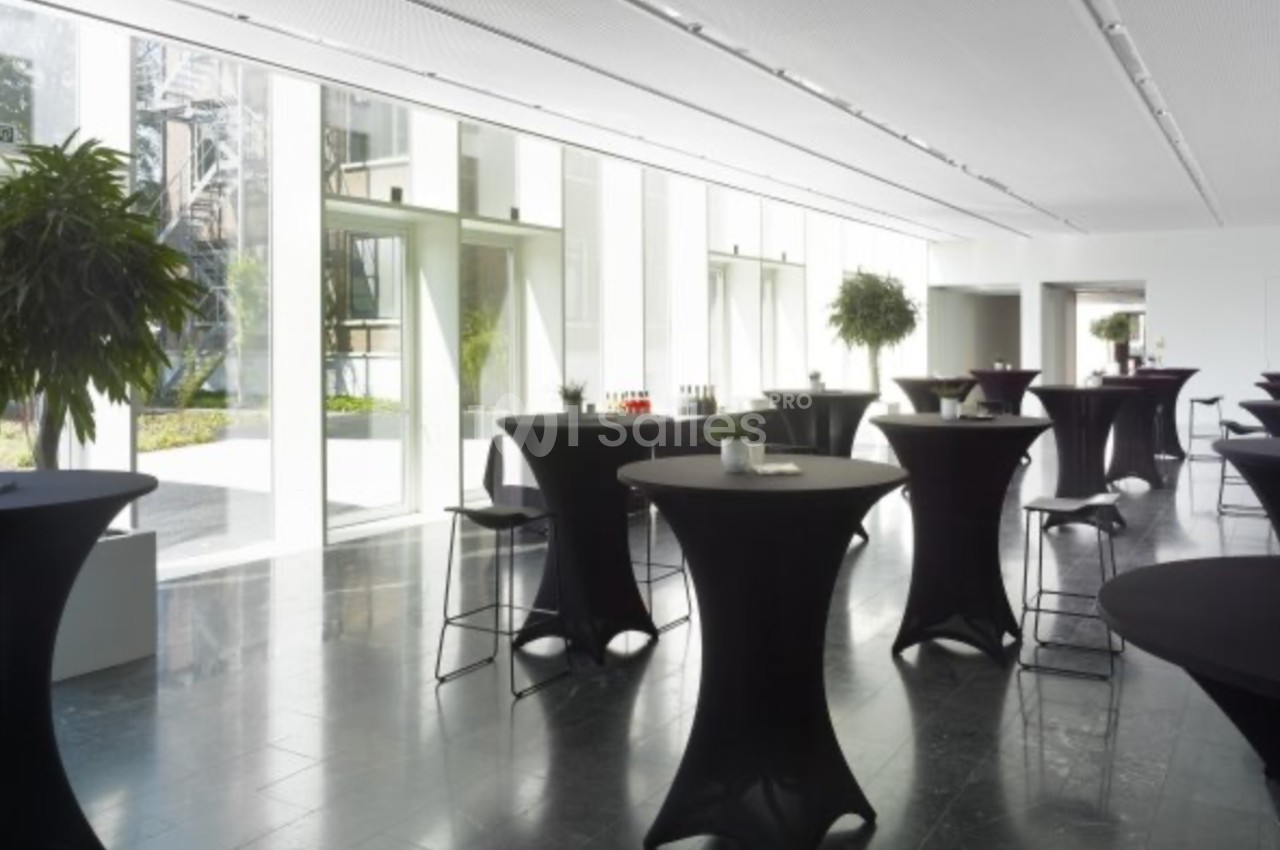 Salle lumineuse avec des tables hautes noires, des plantes en pot et des baies vitrées donnant sur un espace extérieur.