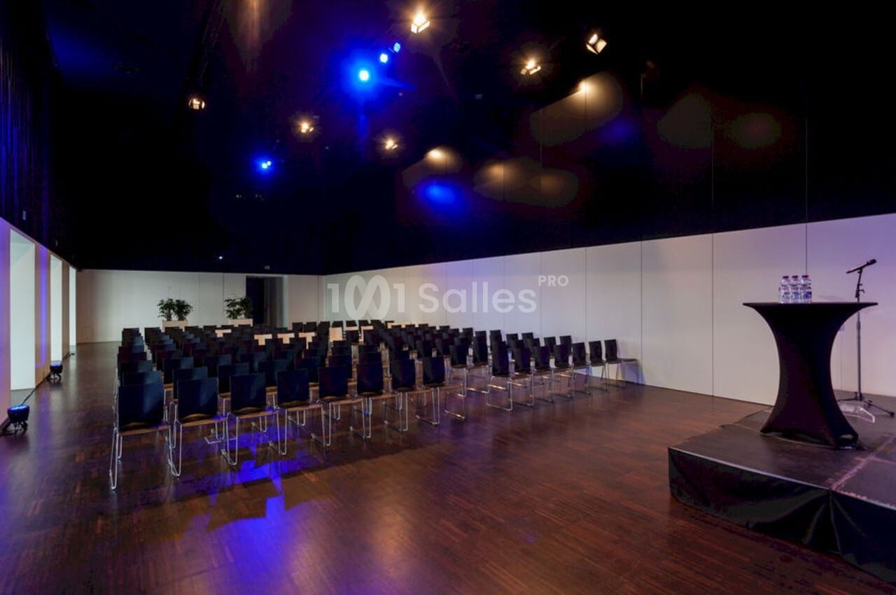 Salle de conférence vide avec rangées de chaises noires, éclairage bleu et une table haute sur une estrade.