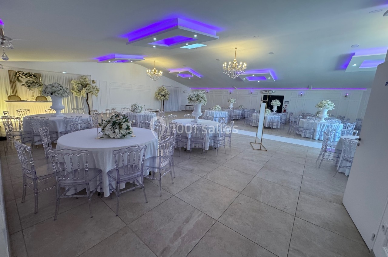 Salle de réception élégante avec tables rondes décorées de fleurs blanches, chaises transparentes et éclairage violet.