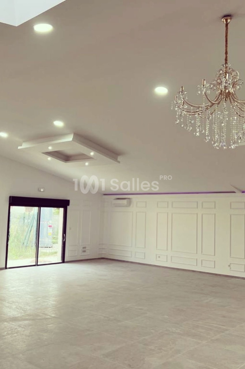 Salle vide avec sol en carrelage, murs blancs ornés de moulures, lustres suspendus et grande baie vitrée donnant sur l…