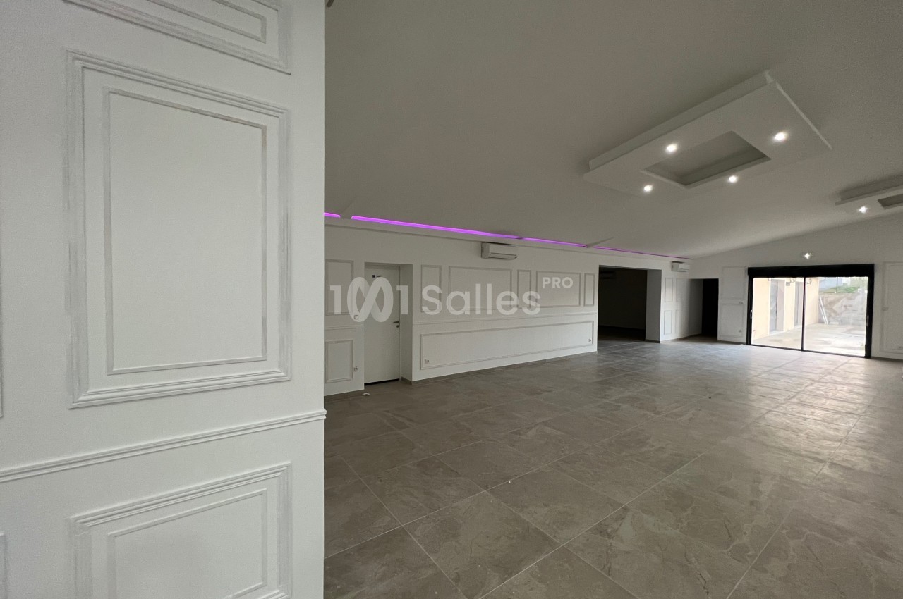 Salle spacieuse avec sol en carrelage, murs blancs moulurés, éclairage encastré et grande ouverture vitrée.