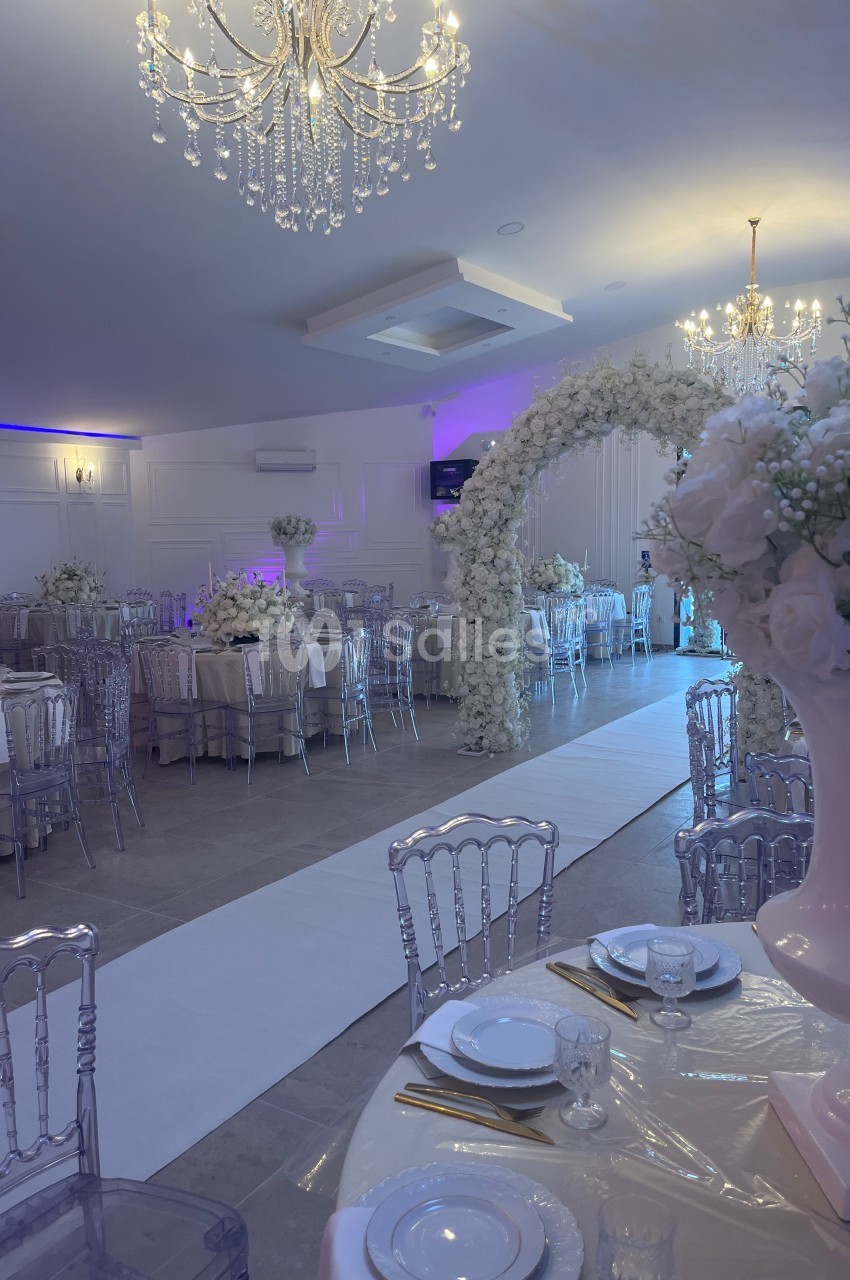 Salle de réception élégante avec tables dressées, chaises transparentes, arche florale blanche et lustres suspendus.