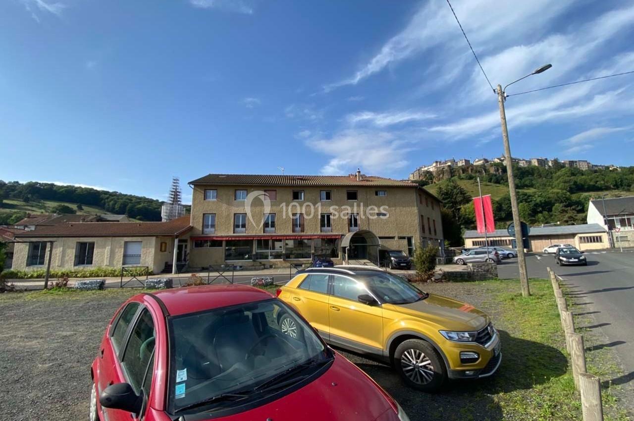 Deux voitures stationnées sur un parking devant un bâtiment en pierre, avec une colline et des maisons en arrière-plan.