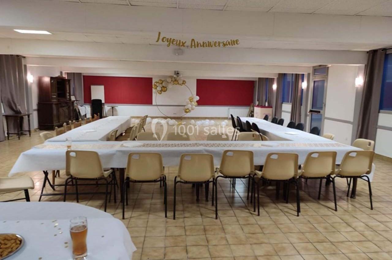Salle décorée pour un anniversaire avec des tables en U, des chaises, des ballons dorés et une banderole ’Joyeux…