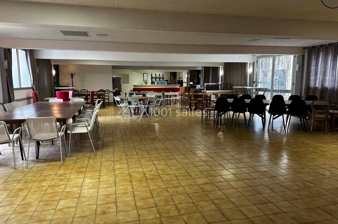 Salle spacieuse avec tables, chaises, bar en arrière-plan et grandes fenêtres laissant entrer la lumière naturelle.