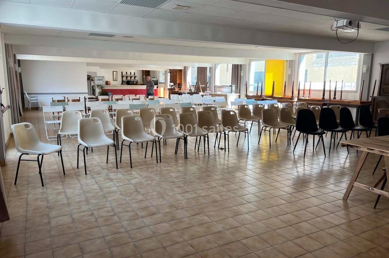 Salle avec des rangées de chaises en plastique alignées, tables empilées et un espace lumineux en arrière-plan.