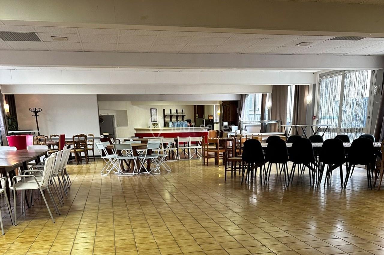 Salle spacieuse avec tables et chaises disposées pour un événement, bar en arrière-plan et lumière naturelle entrant par les…