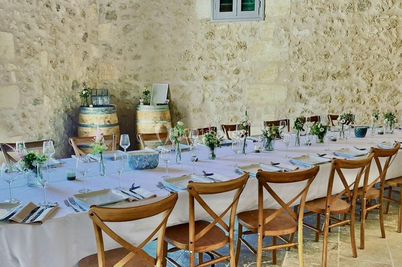 Table dressée avec nappes blanches, chaises en bois, vaisselle élégante et décor floral dans une salle en pierre claire.
