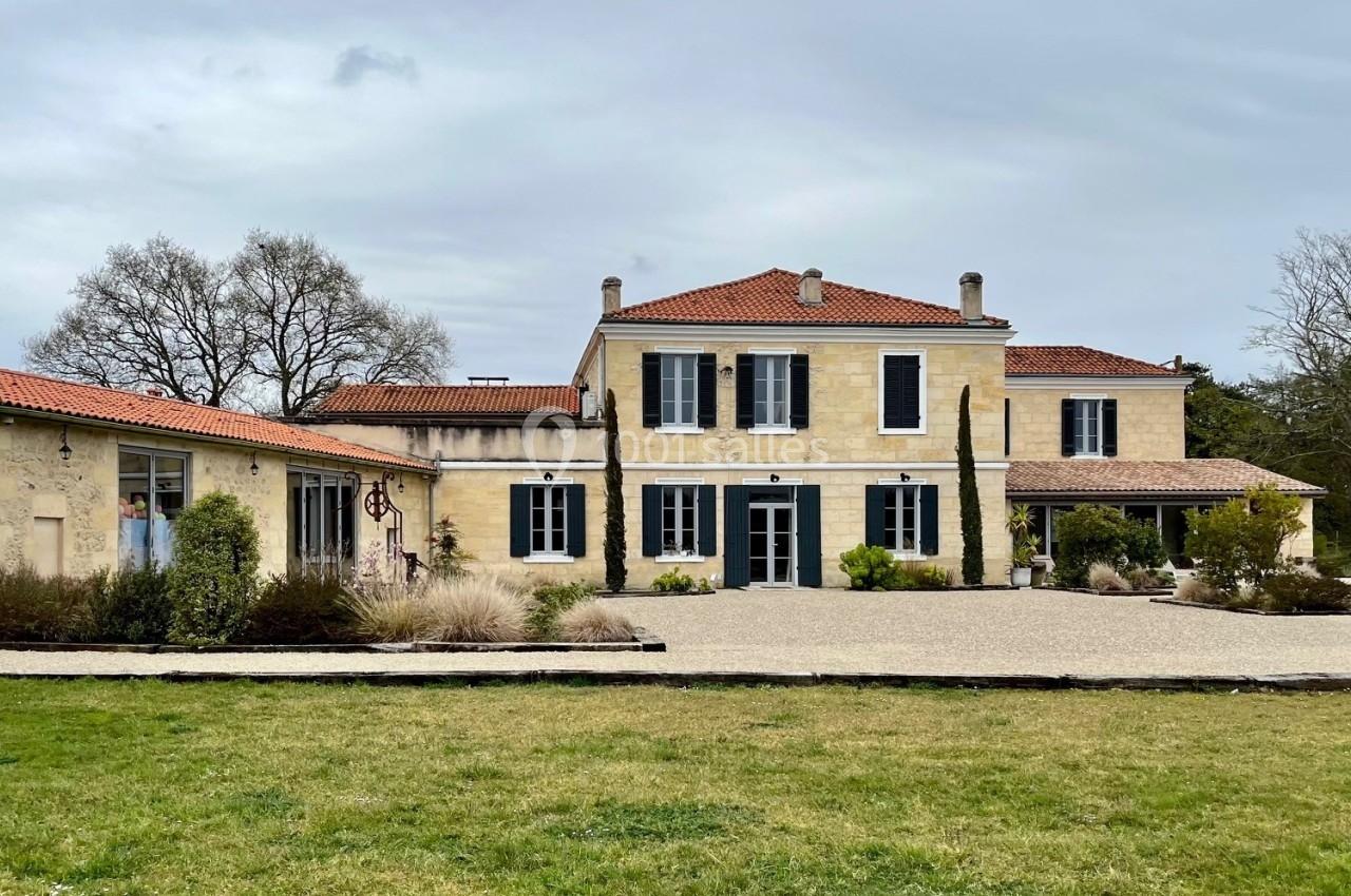 Grande maison en pierre avec volets bleus, entourée d'un jardin et d'une allée gravillonnée, sous un ciel nuageux.