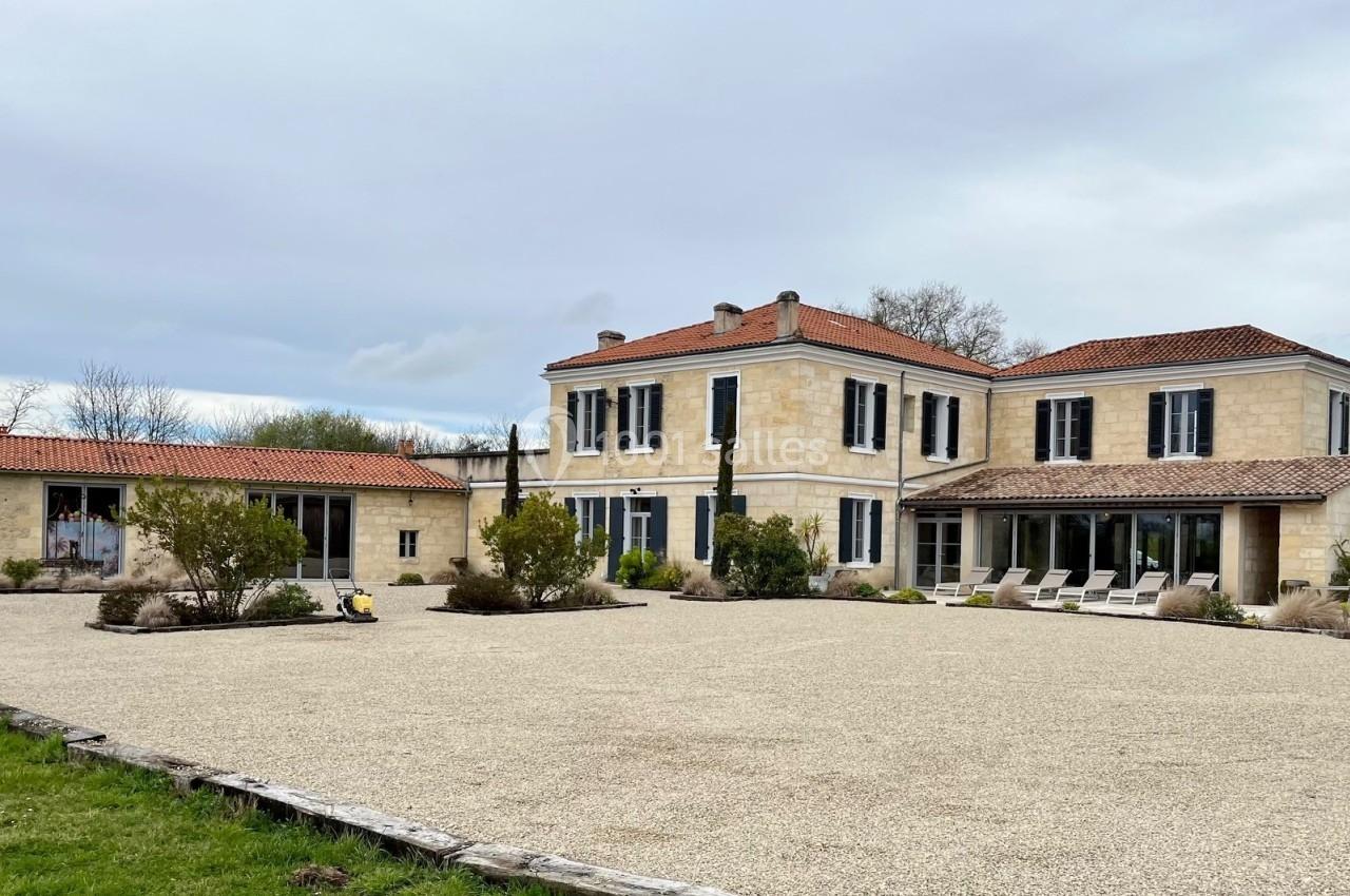 Grande maison en pierre avec volets bleus, toit en tuiles rouges, entourée d'une cour gravillonnée et de végétation.
