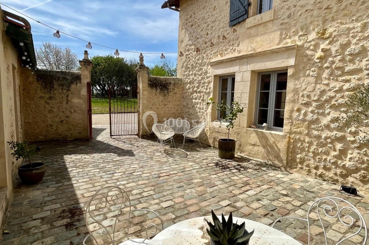Cour pavée ensoleillée avec table blanche, chaises en métal, plantes en pot et façade en pierre d'une maison ancienne.