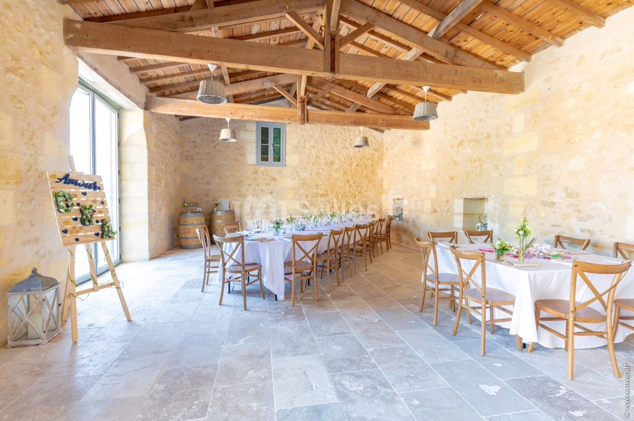 Salle en pierre avec tables rondes et rectangulaires dressées, éclairée par une grande baie vitrée et un plafond en bois.