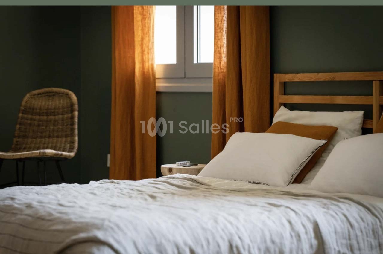 Lit avec draps blancs, coussins et tête de lit en bois, près d'une fenêtre encadrée de rideaux orange, chaise en rotin à…