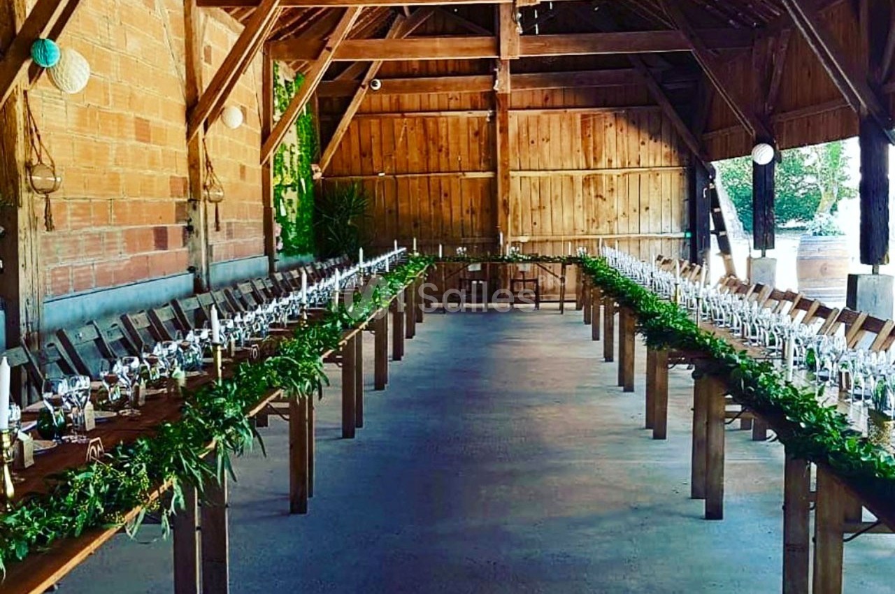 Deux longues tables décorées de verdure et de bougies dans une grange en bois aménagée pour un événement.