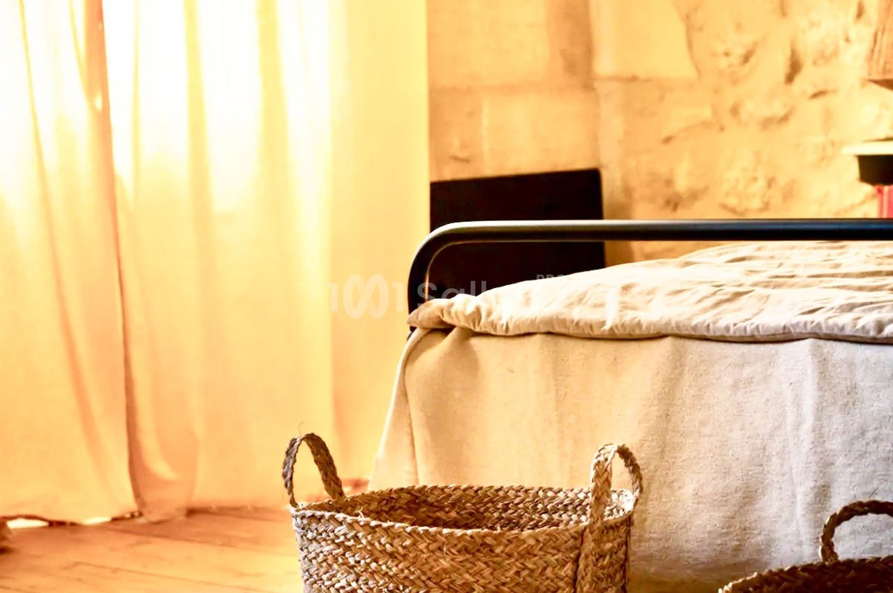 Panier en osier posé près d'un lit avec draps clairs, dans une pièce lumineuse aux murs en pierre.