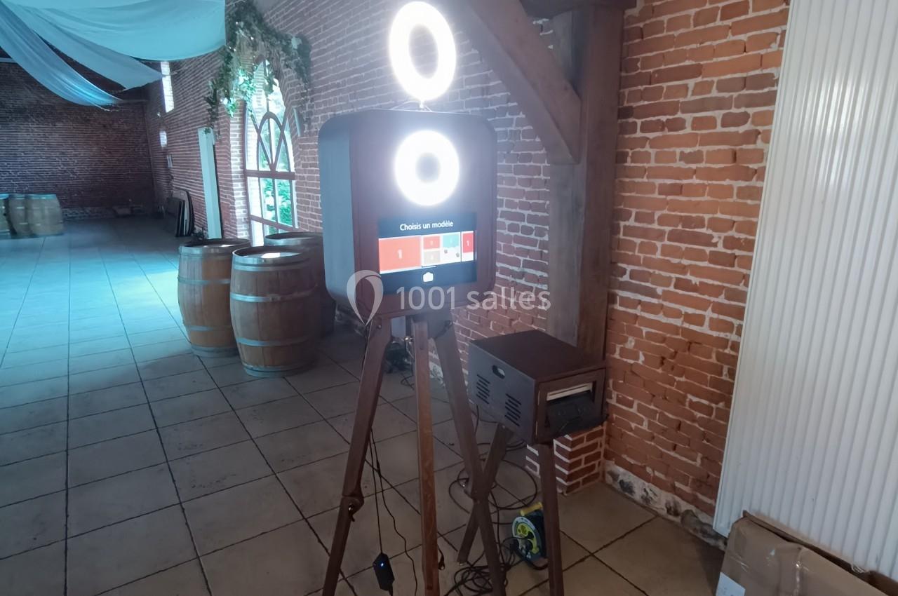 Borne photo sur trépied avec anneaux lumineux, installée dans une salle en briques avec tonneaux en arrière-plan.