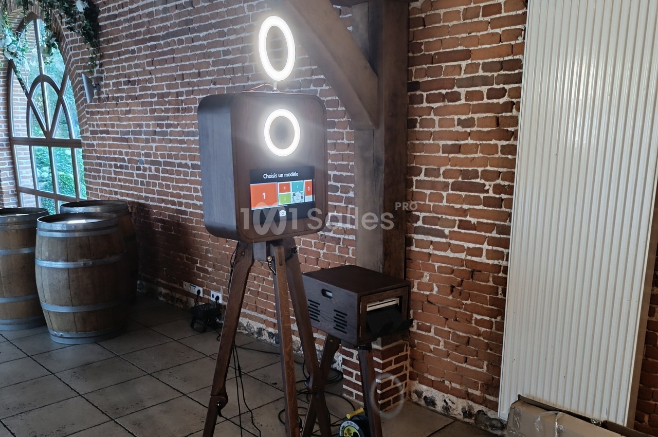 Borne photo sur trépied avec écran tactile et anneaux lumineux, installée dans une salle au mur de briques.