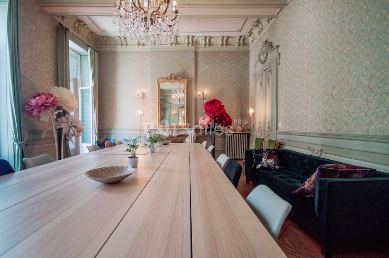 Salle élégante avec une longue table en bois clair, chaises modernes, canapé sombre et décoration florale colorée.