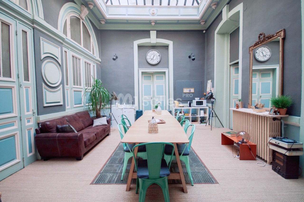 Salle lumineuse avec grande table en bois entourée de chaises vertes, canapé en cuir, plantes et décoration vintage.