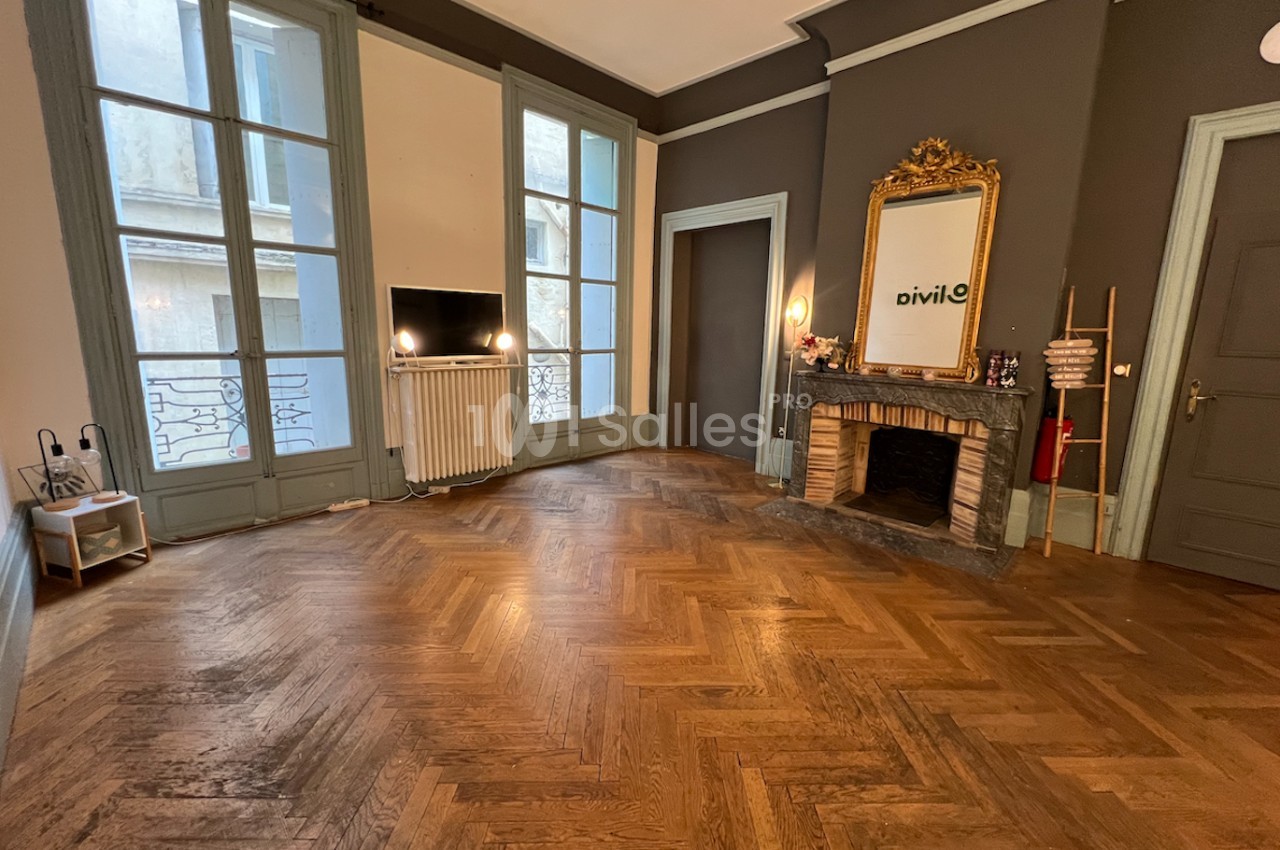 Salon lumineux avec parquet en chevrons, cheminée ornée d'un miroir doré et grandes fenêtres donnant sur l'extérieur.