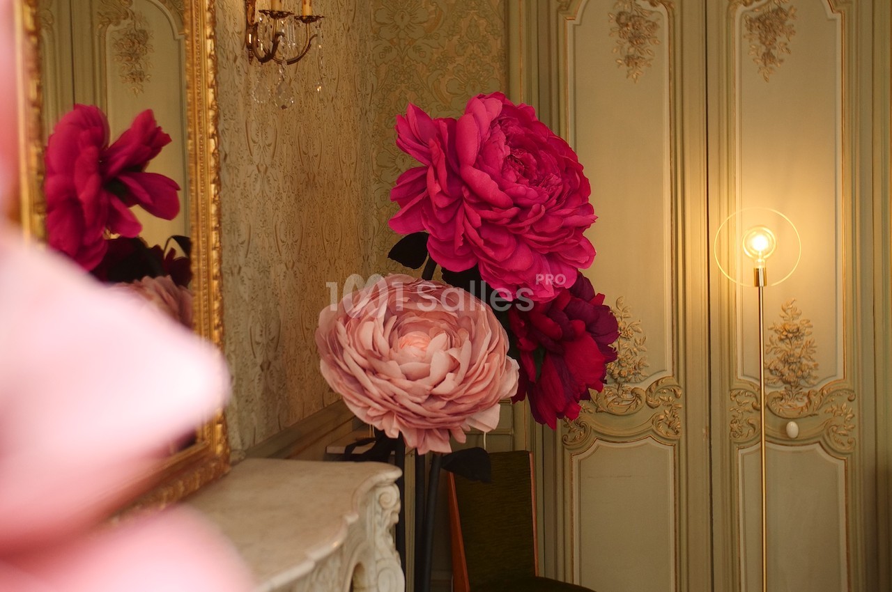 Grandes fleurs décoratives roses et rouges disposées dans une pièce élégante avec moulures et éclairage tamisé.
