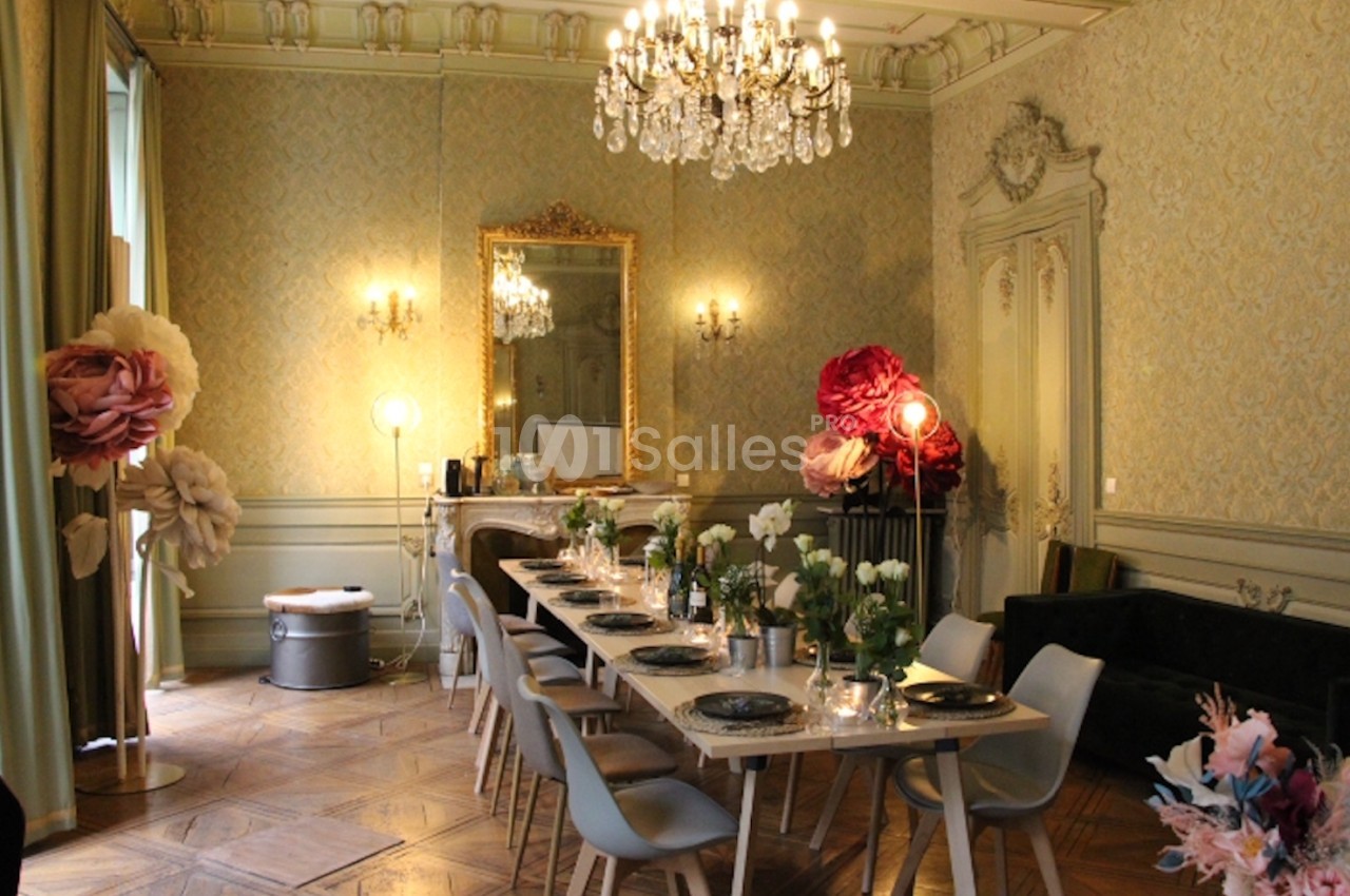 Salle élégante avec une longue table dressée, décorée de fleurs, lustre en cristal et miroir doré sur un mur orné.