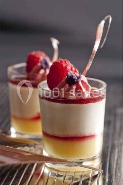 Deux verrines de dessert avec couches de mousse, coulis rouge, framboises et cuillères en métal sur une table.