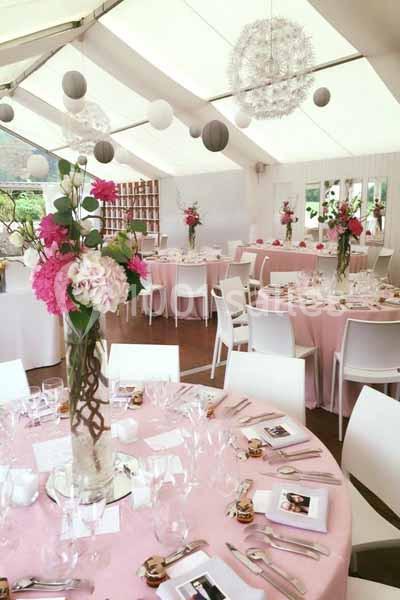 Salle de réception décorée avec des tables rondes nappées de rose, des fleurs en centre de table et des suspensions blanches.