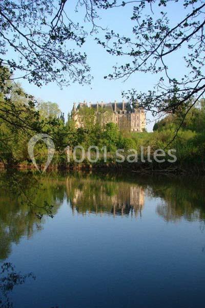 Vue d'un château entouré de verdure, reflété dans un étang calme sous un ciel dégagé.