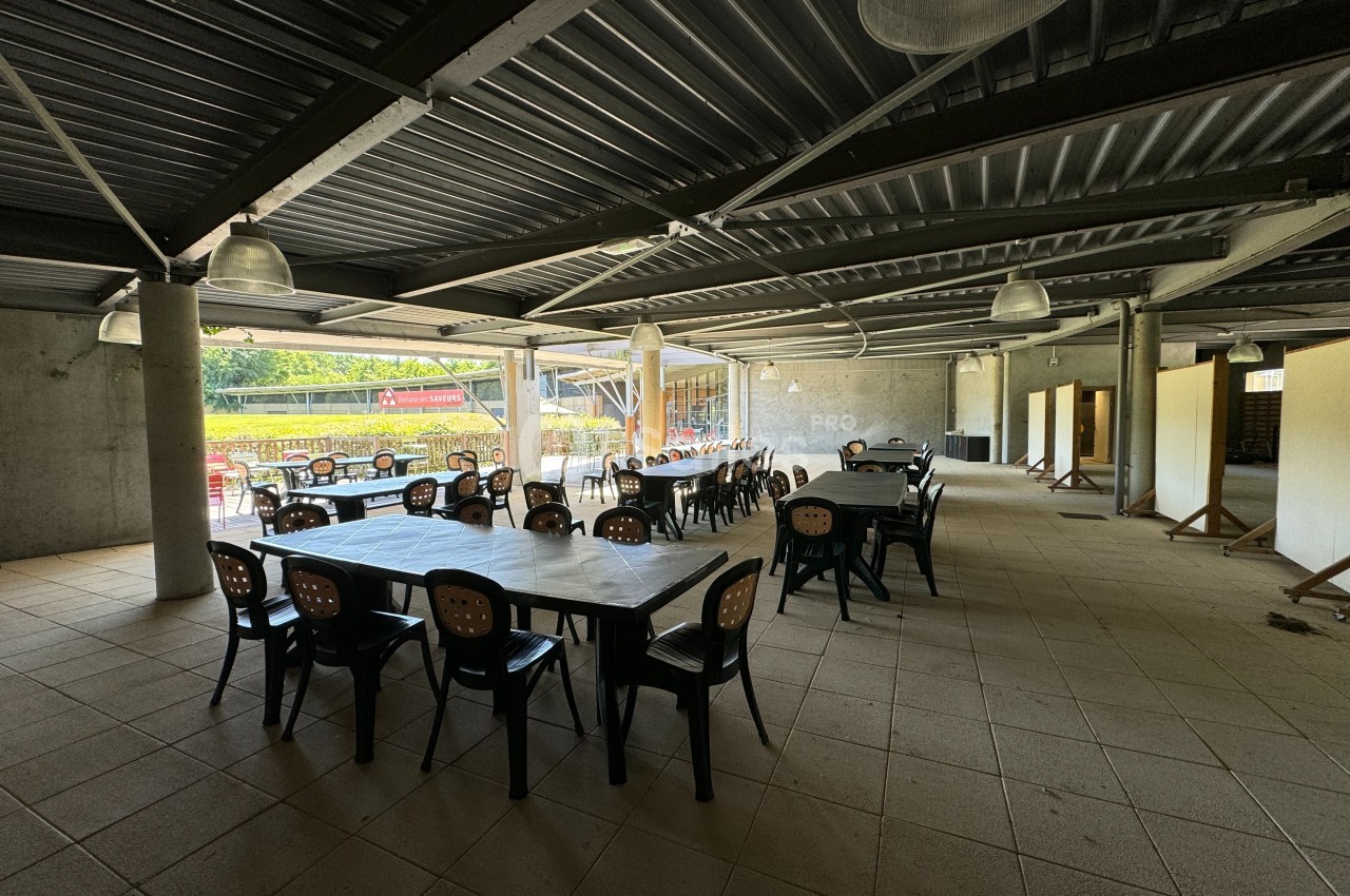 Salle couverte avec des tables et chaises en plastique noir, éclairée par des lampes suspendues et ouverte sur l'extérieur.