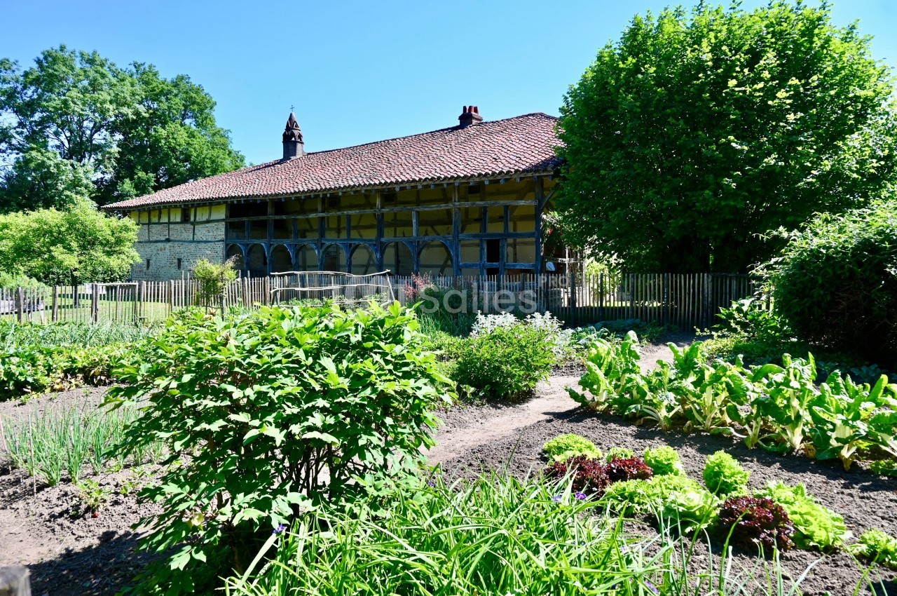 Jardin potager verdoyant devant une maison à colombages avec toit en tuiles, entourée de végétation et d'une clôture en bois.
