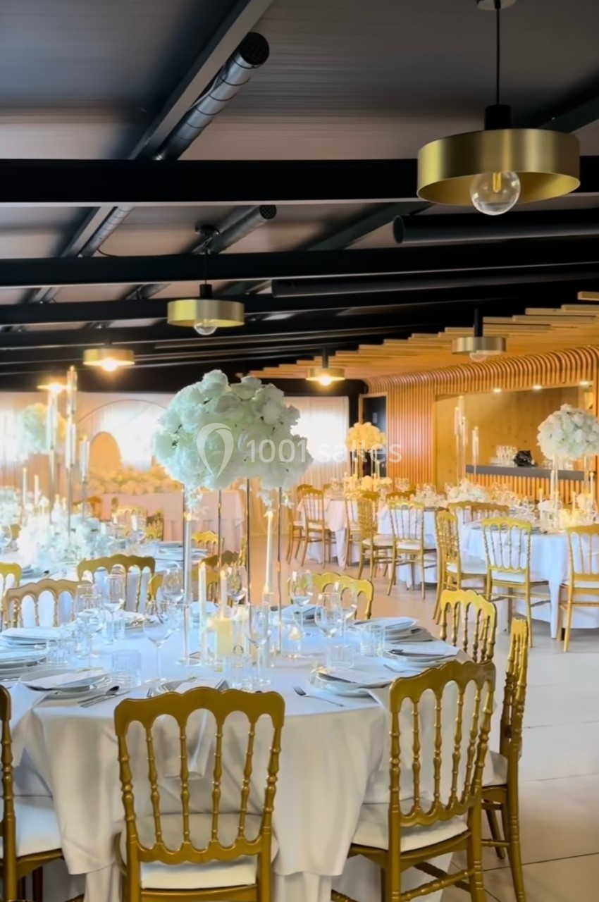 Salle de réception élégante avec tables rondes dressées, chaises dorées et décorations florales blanches.