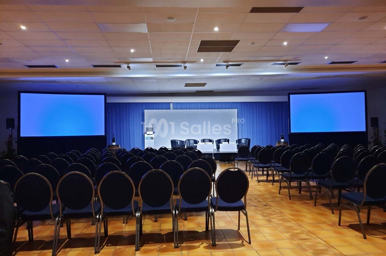 Salle de conférence vide avec des rangées de chaises, un podium et deux écrans de projection allumés.