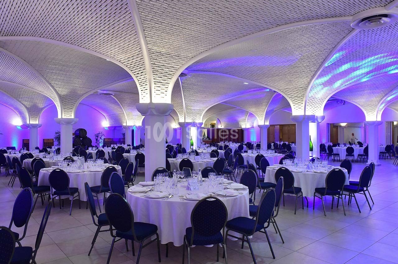 Salle de réception élégante avec tables rondes dressées, chaises bleues et éclairage violet sous des voûtes en pierre.