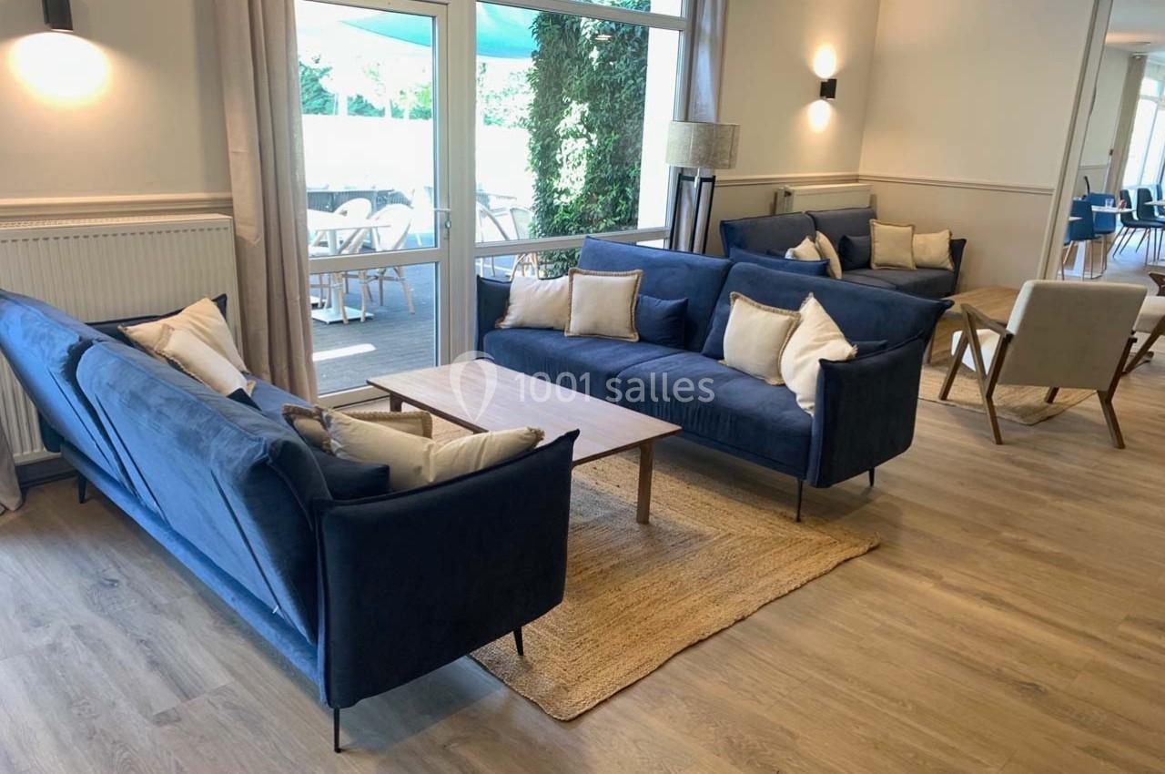 Salon lumineux avec canapés bleus, table basse en bois, tapis beige et vue sur une terrasse extérieure.