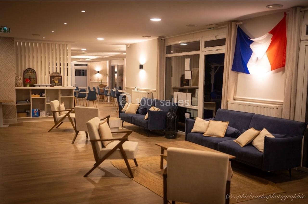 Salon lumineux avec canapés bleus, fauteuils en bois, drapeau français au mur et étagères en arrière-plan.