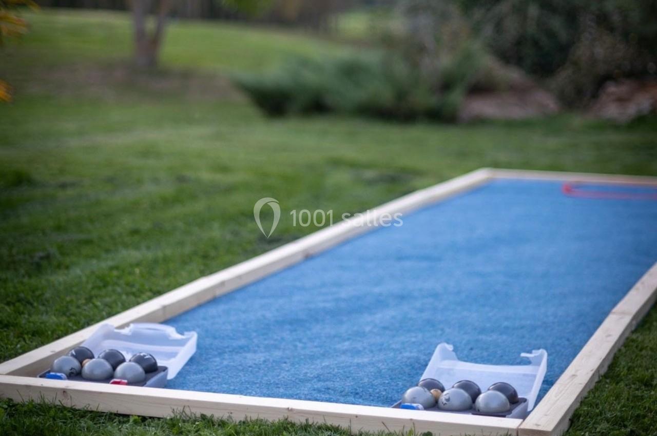 Terrain de pétanque miniature avec tapis bleu, entouré d'herbe, et boules rangées dans des supports aux extrémités.