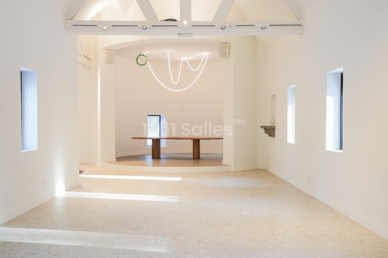 Salle lumineuse avec murs blancs, sol clair, poutres apparentes et une table en bois au fond sous un éclairage minimaliste.