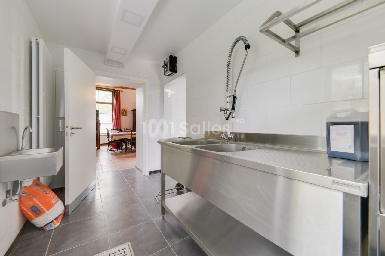 Cuisine professionnelle avec évier en inox, robinet flexible, carrelage gris au sol et vue sur une pièce adjacente.