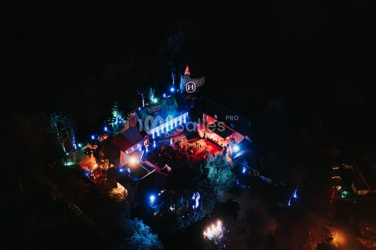 Vue aérienne nocturne d'un bâtiment illuminé par des lumières colorées, entouré de végétation sombre.