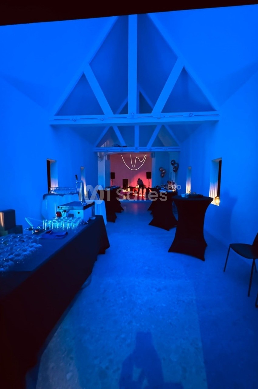Salle éclairée en bleu avec tables dressées pour un événement, et une zone rouge illuminée en arrière-plan.