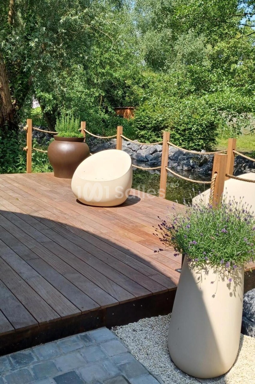 Terrasse en bois avec fauteuils modernes, pots de fleurs et vue sur un jardin verdoyant près d'un cours d'eau.